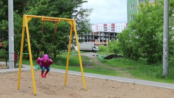 Beetje mooi meisje in regen op schommel op speelplaats in bewolkte dag — Stockvideo