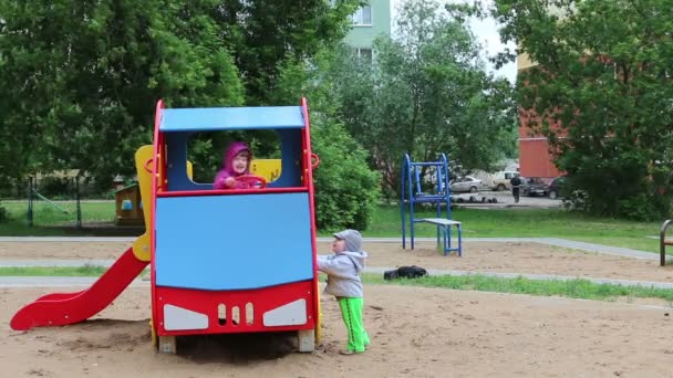 Маленькая счастливая девочка сидит в деревянной машине, а мальчик стоит рядом с ней на детской площадке. — стоковое видео