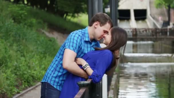 Молодой человек наклоняется над женщиной и целует ее возле маленькой реки в парке в летний день — стоковое видео