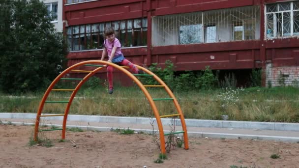 Mała dziewczynka siedzi na półokrągłe drabiny i fale rękę na placu zabaw — Wideo stockowe