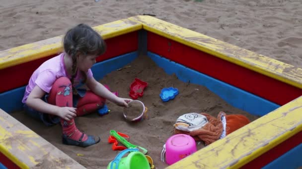 Маленькая милая девочка сидит и играет с игрушками в песочнице на детской площадке — стоковое видео