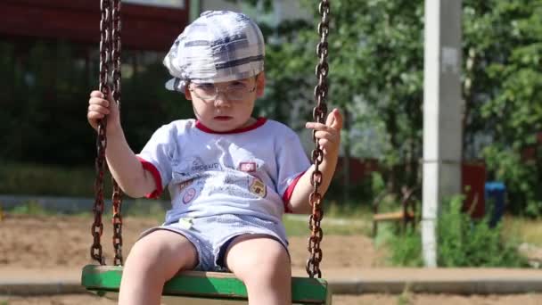 Kleine knappe jongen in glazen swingt op speelplaats op zomerdag — Stockvideo