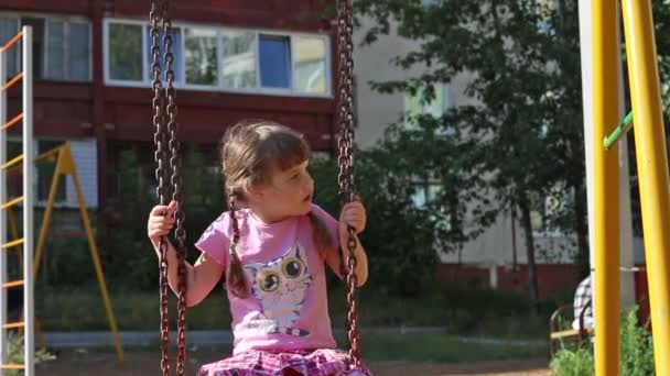 Niña bonita en columpios rosados en el patio de recreo en el día soleado — Vídeo de stock