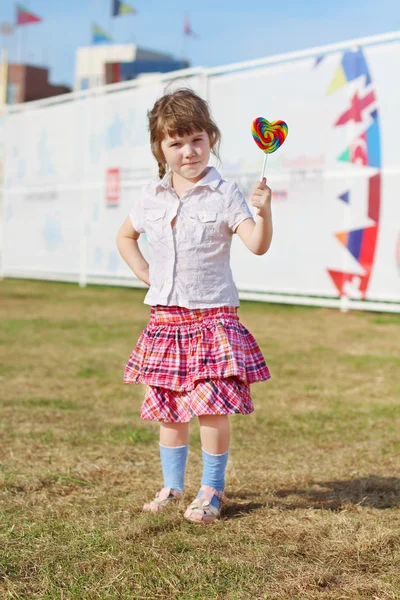 Happy little girl in skirt shows lollipop on grass outdoor — Stock Photo, Image