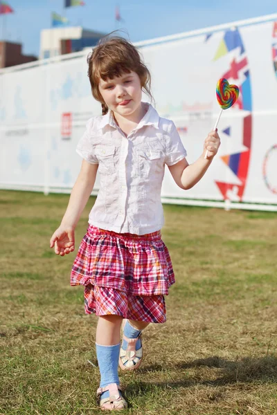 Happy little girl in skirt goes with lollipop on grass — Stock Photo, Image