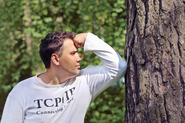 Junger schöner Mann in Weiß lehnt seinen Ellbogen an Baum und denkt — Stockfoto