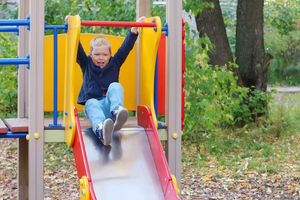 Knappe jongetje speelt op dia op speelplaats in herfstdag — Stockfoto