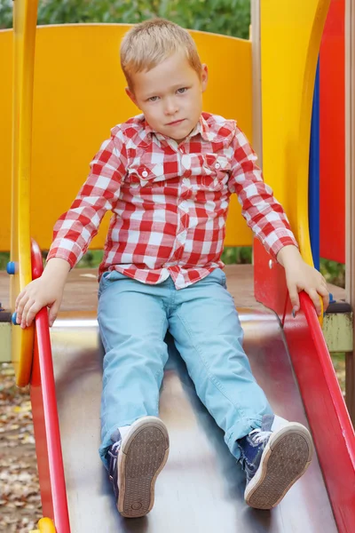 Menino bonito na camisa senta-se no slide no parque infantil — Fotografia de Stock