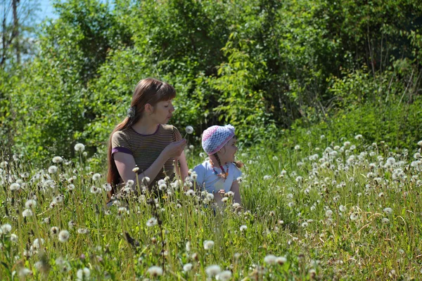 Young mother blows dandelion and little daughter sits near she o — Stock Photo, Image