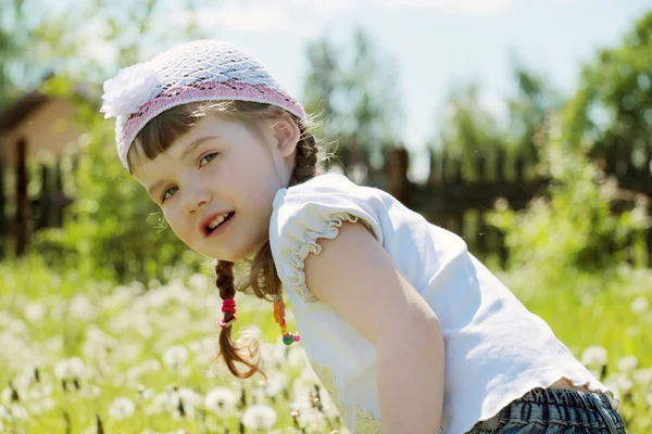 Bella bambina in cappello guarda la fotocamera nel villaggio con dandel — Foto Stock