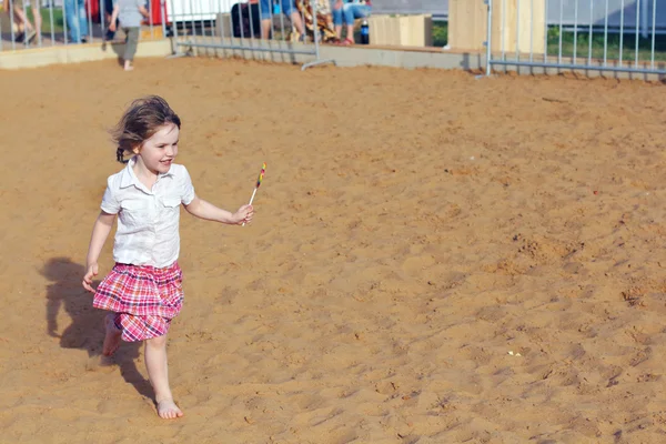 Fröhliches kleines Mädchen läuft im Sommer mit buntem Lutscher auf Sand — Stockfoto