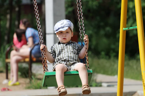 Handsome little boy in cap swings on children playground — Stock Photo, Image