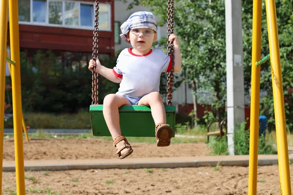 Kleine knappe jongen in cap en glazen swingt op speelplaats — Stockfoto