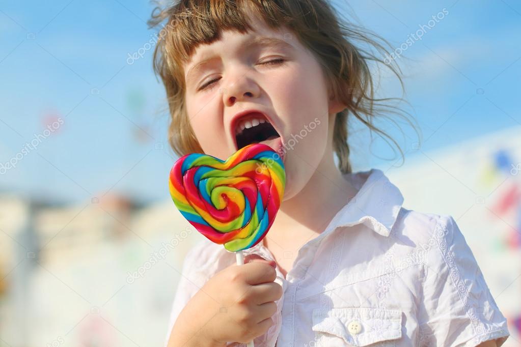 Happy little girl licks bright lollipop outdoor in town