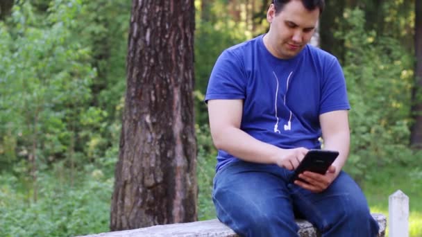 Sonriente hombre se sienta en la pared de piedra y trabaja con la tableta PC en el parque de verano — Vídeo de stock