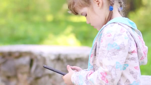 Маленькая девочка сидит на каменной стене и играет с планшетным ПК в парке — стоковое видео