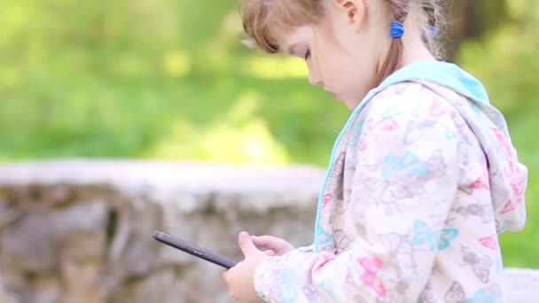 Liten flicka sitter på stenmuren och spelar med tablet Pc i park — Stockvideo