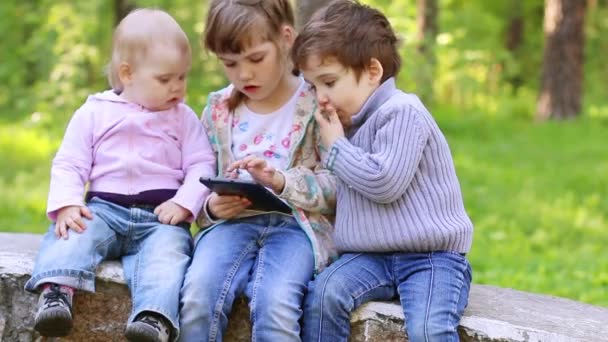 Little girl plays with tablet PC, boy and little girl look at it in summer park — Stock Video