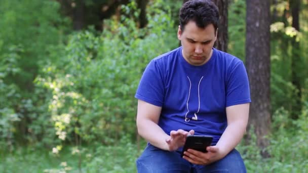 Homem bonito jovem senta-se na parede de pedra e trabalha com tablet PC no parque de verão — Vídeo de Stock