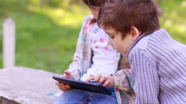 Маленькая девочка играет с планшетным ПК и брат смотрит на него в летнем парке — стоковое видео