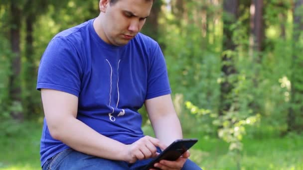 Hombre guapo se sienta en la pared de piedra y trabaja con la tableta PC en el parque de verano — Vídeos de Stock