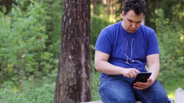 El hombre serio se sienta en la pared de piedra y trabaja con la tableta PC en el parque de verano — Vídeos de Stock