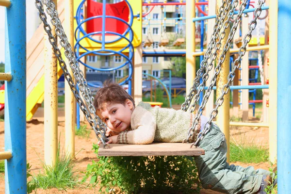 Guapo niño se encuentra en columpios con cadenas en el patio de recreo — Foto de Stock