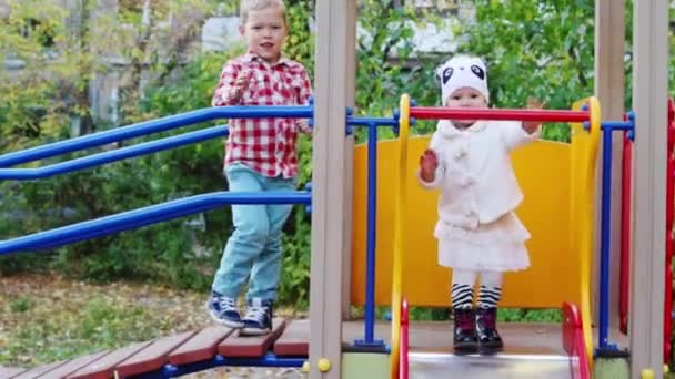 Niño pequeño en camisa a cuadros rojo ondea su mano en el patio de recreo con su hermana — Vídeos de Stock