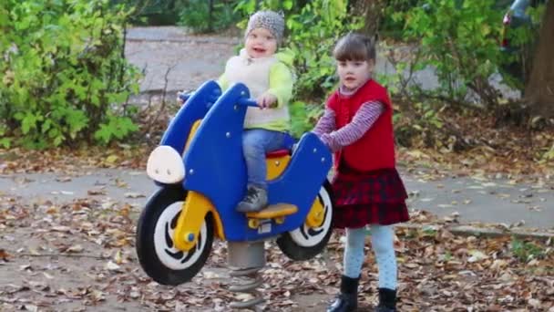 Menina em colete vermelho na moto de madeira jogando no playground com sua irmã mais nova — Vídeo de Stock