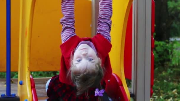 Маленькая девочка в красном жилете висит на руке головой вниз на детской площадке — стоковое видео