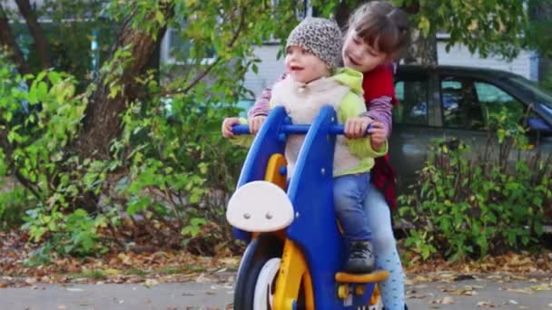 Menina em colete vermelho em moto de madeira jogando no playground com sua irmã mais nova — Vídeo de Stock