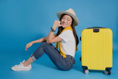 Günlük giysiler içinde Asyalı kadın turist mavi arka planda izole bir şekilde bavulun yanında oturuyor. Bavuluna işaret etti..