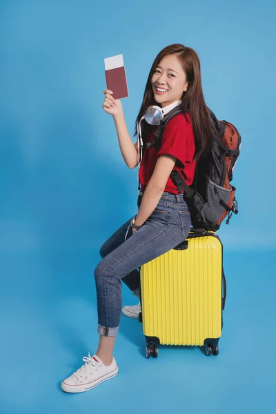 Young Asian tourists are smiling and excited for their journey. She is sitting on a yellow suitcase with a passport on a blue background in the studio.