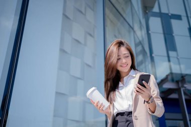 Profesyonel Asyalı iş kadını kahve içiyor ve dışarıda dolaşırken internetten kitap okuyor. Ofisinin yanında.