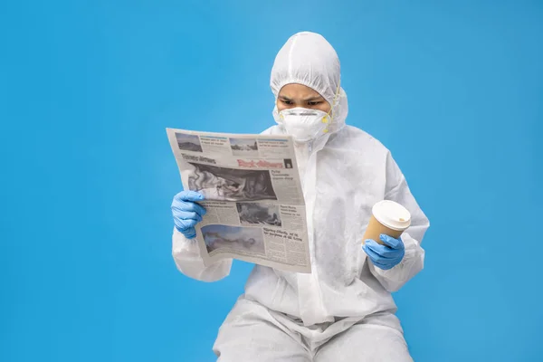 手袋とマスクで保護スーツのアジアの医師 彼女は真剣に手に新聞とホットコーヒーを読んでいます — ストック写真