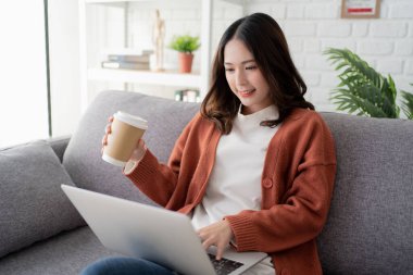 Gülümseyen genç bir kadın sabah kahvesinden zevk alıyor ve dizüstü bilgisayar kullanıyor. Evde oturup çalışıyor..