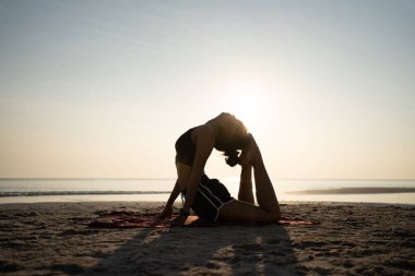 Gün batımında yoga yapan Asyalı kadın silueti. Sahilde yoga pozu vermek için sırtını kavis yapıp ayaklarıyla başını okşuyor. Sağlık hizmeti kavramı.