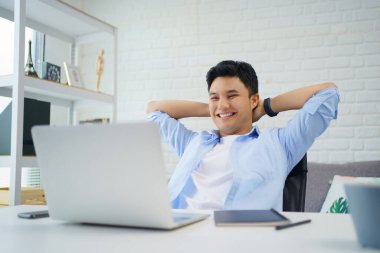 Genç Asyalı adam gülümsüyor ve evde çalışırken bilgisayarından ekranı okuyor..