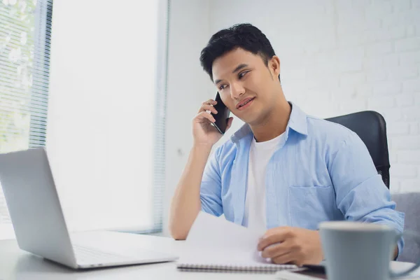 Young Asian businessman talks on the phone and watches his laptop during the workflow at work.