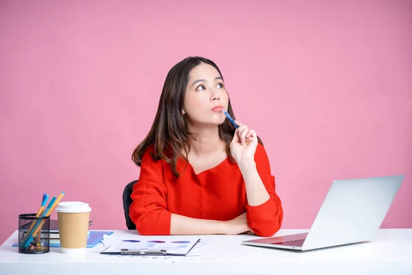 Young Asian woman was thinking about something with holds a pen near her chin and looks up at her desk. Isolated pastel pink background.