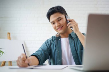 Young Asian man in casual clothes is taking notes while talking on the phone on the desk in the room.