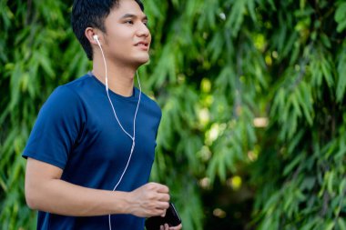 Yakışıklı Asyalı adam akıllı telefondan müzik dinliyor ve parkta koşuyor..