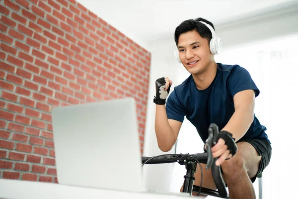 トレーナーに自転車に乗ってオンラインで自転車ゲームをして家の中で運動しているアジア人男性は 彼が満足していた — ストック写真