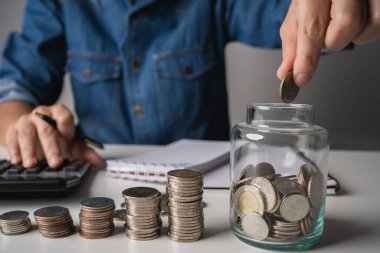 Businessman hand holding coins putting in jar and recording income and expenses in the account book, investments for future.