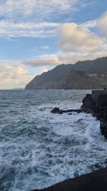 Madeira 'daki Porto da Cruz Körfezi' nde kıyı şeridi boyunca doğal deniz havuzları ve güçlü dalga hareketleriyle dalgalı bir Atlantik Denizi yer alıyor. Sahne, suyun volkanik kaya oluşumlarına karşı enerjik hareketini yakalar ve ham kıyı bölgesini vurgular.