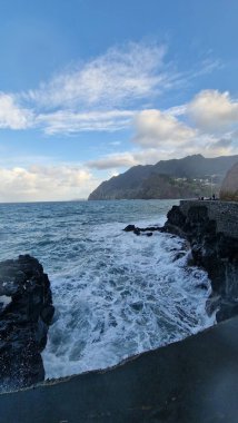 Madeira 'daki Porto da Cruz Körfezi' nde kıyı şeridi boyunca doğal deniz havuzları ve güçlü dalga hareketleriyle dalgalı bir Atlantik Denizi yer alıyor. Sahne, suyun volkanik kaya oluşumlarına karşı enerjik hareketini yakalar ve ham kıyı bölgesini vurgular.