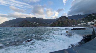 Madeira 'daki Porto da Cruz Körfezi' nde kıyı şeridi boyunca doğal deniz havuzları ve güçlü dalga hareketleriyle dalgalı bir Atlantik Denizi yer alıyor. Sahne, suyun volkanik kaya oluşumlarına karşı enerjik hareketini yakalar ve ham kıyı bölgesini vurgular.