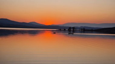 Milada Gölü 'nün üzerinde sihirli turuncu bir günbatımı. Gökyüzü sıcak renklerle parlıyor ve durgun suya yansıyor. Huzurlu göl, parlayan bulutları yansıtır. Huzurlu ve büyüleyici bir atmosfer yaratır. Alacakaranlıktaki doğanın güzelliğini ön plana çıkarır..