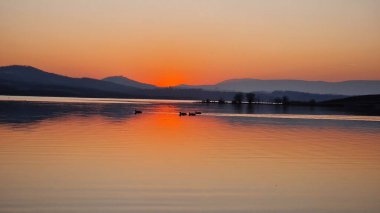 Milada Gölü 'nün üzerinde sihirli turuncu bir günbatımı. Gökyüzü sıcak renklerle parlıyor ve durgun suya yansıyor. Huzurlu göl, parlayan bulutları yansıtır. Huzurlu ve büyüleyici bir atmosfer yaratır. Alacakaranlıktaki doğanın güzelliğini ön plana çıkarır..