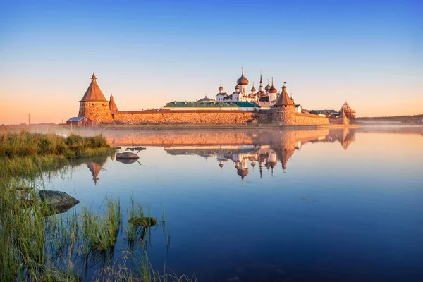 ソロヴェツキー修道院の鏡像ソロヴェツキー島の聖なる湖の穏やかな水の中で夜明けの太陽と海岸近くの草の光の中で — ストック写真